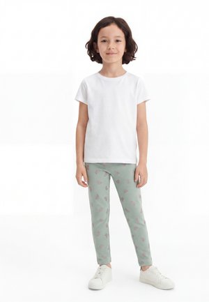 Copil cu păr închis la culoare până la umeri, purtând un tricou alb simplu, pantaloni verzi cu modele roz și adidași albi, în picioare cu fața înainte.