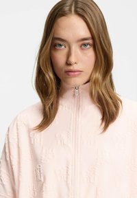 Chaqueta rosa claro con cremallera y un patrón texturizado que muestra el nombre de la marca. Cuello alto y tejido suave que crean un diseño contemporáneo y relajado.