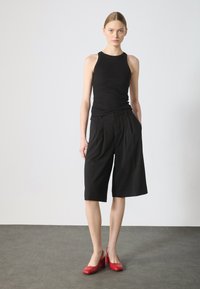 Top in tessuto a coste nera abbinato a pantaloni corti neri plissettati fino al ginocchio e scarpe con tacco rosse. Lo sfondo è semplice e di colore chiaro.
