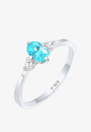 Anillo de plata de ley que presenta una gema azul ovalada con dos pequeñas piedras transparentes a cada lado, destacando un diseño elegante y pulido.
