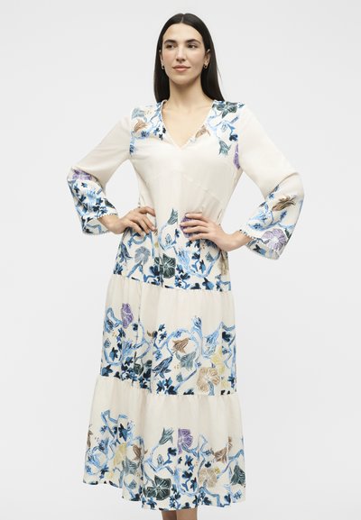 Femme aux longs cheveux foncés portant une robe midi crème avec des motifs floraux bleus, violets et bruns, debout les mains sur les hanches.