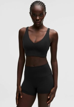 Donna con capelli intrecciati che indossa un reggiseno sportivo nero e pantaloncini a vita alta abbinati, in piedi contro uno sfondo chiaro e uniforme.