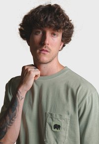 Olivově zelené tričko z bavlněného jerseye, s přišitým kapsou a černým logem slona. Model předvádí volný střih a kulatý výstřih.