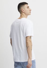 Camiseta blanca de algodón con mangas cortas, cuello redondo y corte relajado. Tatuajes visibles en el brazo. El modelo está de perfil, girado ligeramente hacia atrás.