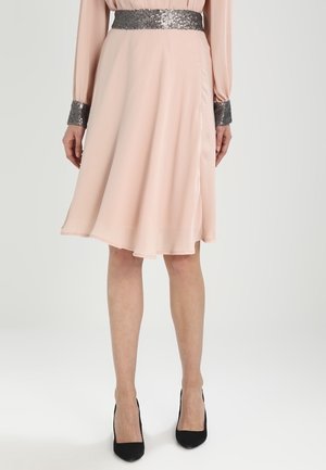 Femme portant une jupe rose pâle longueur genou avec une ceinture et des poignets ornés de sequins argentés, assortie à des chaussures à talons noirs.