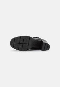 Semelle de chaussure noire en caoutchouc texturé, dotée de rainures horizontales et de six crampons de traction, conçue pour la stabilité et la durabilité.