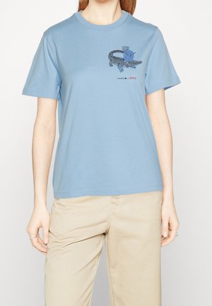 Person wearing a light blue T-shirt with a graphic of a crocodile carrying a blue bird, paired with beige pants.