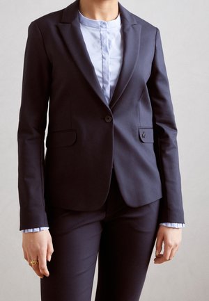 Blazer bleu marine ajusté avec deux poches avant, fermeture à un bouton et revers crantés. Chemise col bleu clair visible en dessous.
