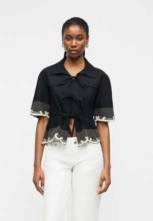 Kvinde iført en sort kortærmet bluse med hvid broderi og snore, kombineret med højtaljede hvide bukser, stående mod en ensfarvet baggrund.