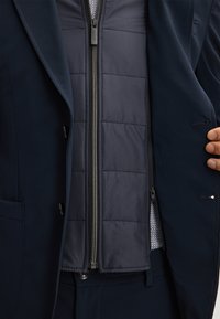 Marineblauw colbert met een ritssluiting en een gewatteerd vest aan de binnenkant. Het vest heeft een gladde textuur en beschikt over kwadranten voor isolatie.