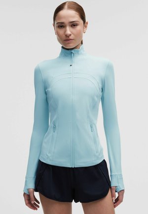 Frau mit roten Haaren, die eine hellblaue, vollverschlossene Sportjacke und schwarze Laufshorts trägt, steht vor einem weißen Hintergrund.