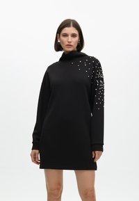 Schwarzes Sweatshirt-Kleid mit hohem Kragen, langen Ärmeln und verstreuten Perlenverzierungen auf einer Schulter. Glattes Material, lockerer Schnitt.