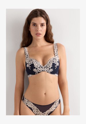 Vrouw met lang bruin haar draagt een marineblauw lingeriesetje met beige bloemenkant, staand tegen een effen lichte muur, naar voren kijkend.