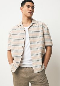 Camicia beige a maniche corte con righe orizzontali blu e crema, tessuto strutturato e bottoni neri, indossata sopra una canottiera bianca.