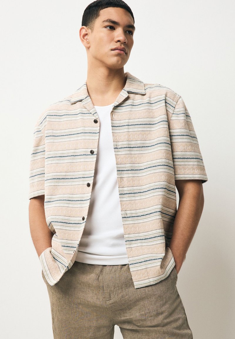 Camicia beige a maniche corte con righe orizzontali blu e crema, tessuto strutturato e bottoni neri, indossata sopra una canottiera bianca.