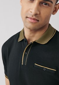 Juoda polo marškinėliai su auksine tekstūruota apykakle, trimis sagomis priekyje ir maža krūtinės kišenė su kontrastine apdaila.