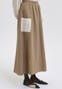 Maxi nederdel i tan-stof med elastisk talje, udstyret med en stor off-white lomme. Sammen med flade sko i sort og creme.