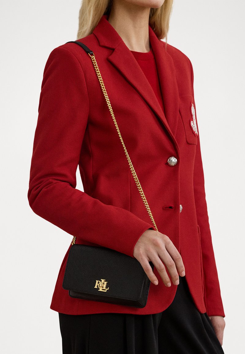 Schwarze, strukturierte Handtasche mit goldenem Logo, als Umhängetasche getragen. Kombiniert mit einem roten Blazer mit silbernen Knöpfen und einer dekorativen Taschenverzierung.