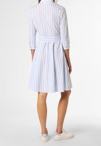 Kvinna klädd i en ljusblå och vit randig knälång klänning med trekvartslånga ärmar och vita sneakers, stående med ryggen mot kameran.