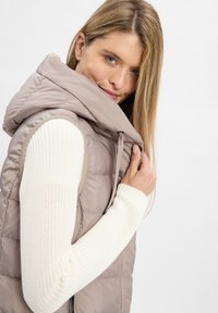 Gepolsterte ärmellose Weste in Hellbeige mit hohem Kragen und Kapuze, kombiniert mit einem strukturierten weißen Langarmshirt. Glatte, weiche Oberfläche.