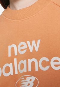 Felpa arancione con colletto a coste. Presenta una grande scritta bianca che dice "new balance" e un logo rotondo sottostante. Tessuto morbido.