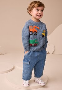 Mosolygós kisgyermek kék pulóvert visel, amelyen színes építőipari járművek vannak, farmer cargo nadrágot és fehér tornacipőt, kezét a zsebében tartva áll.