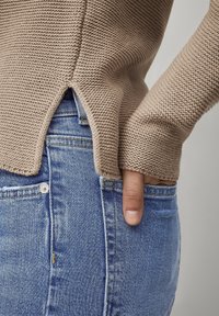 Beige gebreide trui met ribbeltextuur, zijsplitdetails, gedragen over lichtblauwe denim jeans met een voorkeurszak en knoopsluiting.