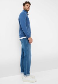 Giacca in pile blu con colletto alto, abbinata a jeans azzurri e sneakers bianche. I jeans hanno un leggero risvolto alle caviglie.