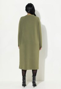 Vestito lungo verde oliva a maniche lunghe con silhouette dritta, tessuto texturizzato e scollatura rotonda, abbinato a stivali neri alla caviglia.