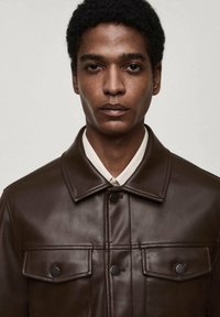 Veste en cuir marron avec un devant boutonné, deux poches poitrine et un col. Texture lisse et coupe ajustée.