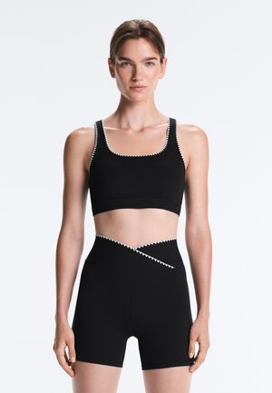 Femme portant un soutien-gorge de sport noir et un short cycliste noir taille haute avec un bord festonné blanc, debout sur un fond blanc uni.