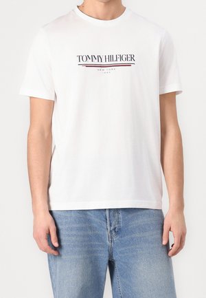 Mand iført en hvid Tommy Hilfiger T-shirt med logo og teksten "New York 1985," kombineret med lyseblå jeans mod en ensfarvet baggrund.