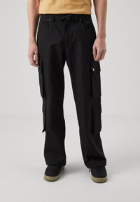 Svarte cargo-bukser med flere lommer, teksturert stoff, justerbar midje og avslappet passform. Brukt med mørke sneakers.