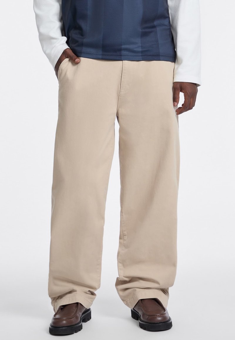Beige wide-leg trousers made of lightweight fabric, featuring side pockets and a straight cut, paired with dark brown shoes.