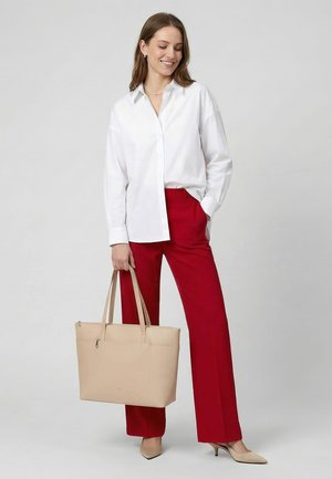 Kvinde iført hvid bluse og røde bukser holder en beige mulepose og smiler med den ene hånd i lommen mod en ensfarvet baggrund.