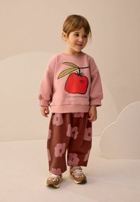 Ružová mikina s grafikou čerešňovej paradajky, skombinovaná s voľnými hnedými nohavicami s ružovými kvetinovými vzormi. Outfit dopĺňajú vzorované tenisky.