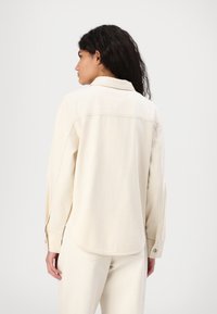 Lys beige denimskjorte med spiss krage, lange ermer og avrundet nederkant. Har knappemansjetter og subtile sømdetaljer.