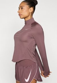 Μακρυμάνικο τοπ με φερμουάρ στο χρώμα μαβί, με ψηλό γιακά, απαλή υφή και λογότυπο Nike. Συνδυάζεται με αντίστοιχα μαβί σορτς για τρέξιμο.