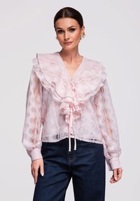 Femme portant un chemisier en dentelle rose transparente avec un grand col volanté et un nœud, associé à un jean bleu foncé et des boucles d'oreilles fleuries.