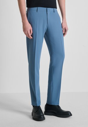 Hombre con pantalones ajustados azul y zapatos de vestir de cuero negro, de pie contra un fondo liso.