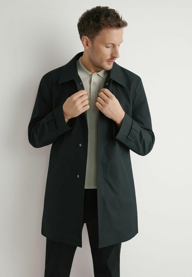 Mężczyzna poprawiający kołnierz czarnego płaszcza na beżowej koszuli zapinanej na guziki, stojący na tle gładkiej, jasnej ściany.