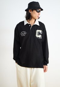 Black long-sleeve polo shirt with a white collar, featuring embroidered details and a large "C" emblem on the chest. Worn with cream pants.