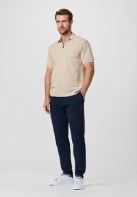 Polo beige con cerniera, dettagli bianchi su colletto e maniche, abbinata a pantaloni della tuta blu navy con strisce laterali e scarpe bianche.