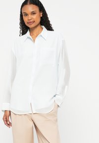 Light blue, long-sleeve button-up shirt with a collar, front pocket, and a relaxed fit. Soft, smooth texture; paired with beige pants.