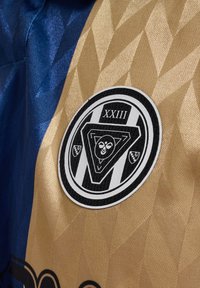 Trøje lavet af struktureret stof i blå og guld med et geometrisk mønster, med et cirkulært sort og hvidt emblem med "XXIII."