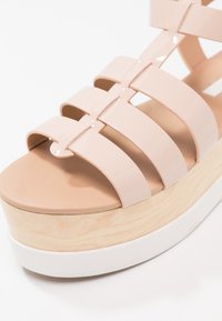Sandale à plateforme beige avec plusieurs larges brides et une semelle intermédiaire en bois, dotée d'une semelle extérieure en caoutchouc blanc sur fond blanc.