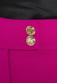 Close-up van een magenta tailleband met twee glanzende gouden decoratieve knopen op een zwarte stoffen achtergrond.