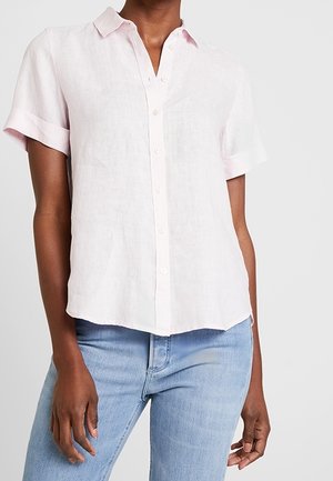 Frau trägt ein hellrosa kurzärmeliges Hemd mit Knöpfen und hellblaue Jeans, steht vor einem schlichten Hintergrund.