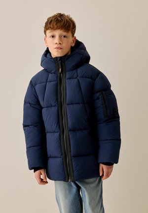 Dreng iført en mørkeblå polstret hættejakke med lynlås foran og lyseblå jeans mod en ensfarvet beige baggrund.