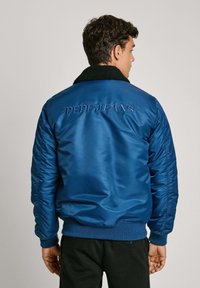 Cazadora bomber de satén azul con un cuello de forro polar negro. Presenta la marca bordada "PEPE JEANS" en la parte posterior y puños y dobladillo acanalados.